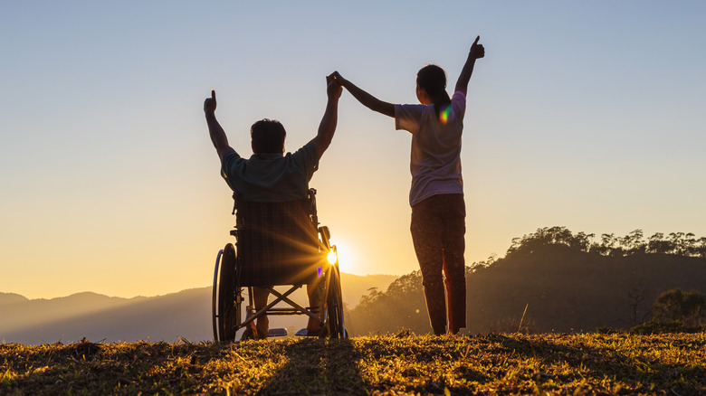 man in wheelchair and woman holding hands up triumphantly against a sunset