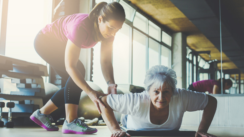 Personal trainer helping an elderly woman do push-ups on an exercise mat.