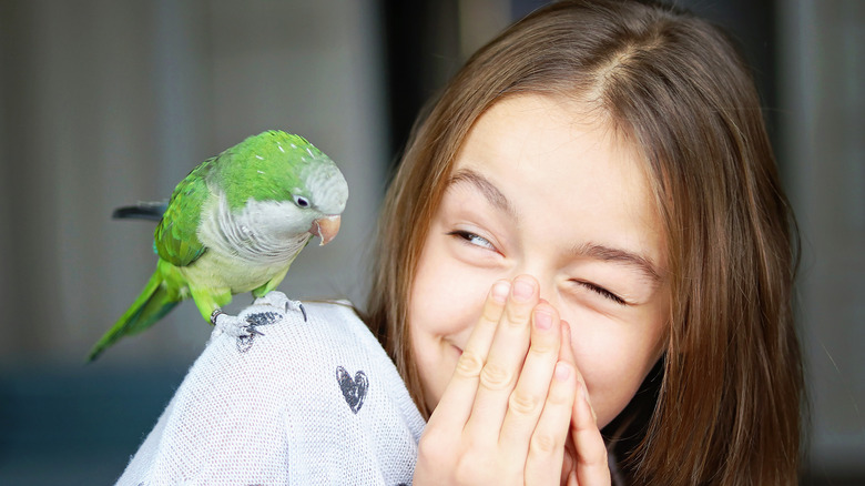 A girl smiling at a bird on her shoulder while covering her nose with her hands