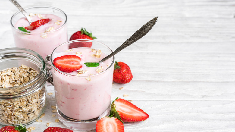 Two glasses of strawberry yogurt surrounded by strawberries and next to a glass jar of granola