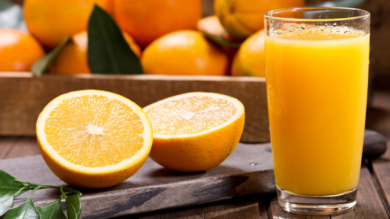 A glass of orange juice next to a halved orange on a cutting board with a square container of oranges in the background