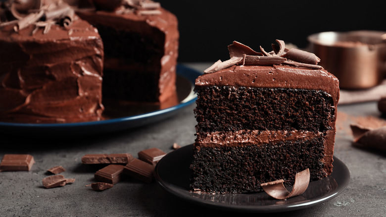 A slice of chocolate layer cake with chocolate icing and topped with chocolate shavings in the foreground with the rest of the chocolate cake in the background