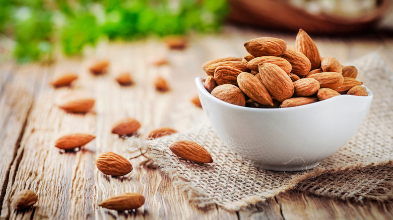 A white bowl filled with almonds on top of burlap and next to scattered almonds on a wooden table