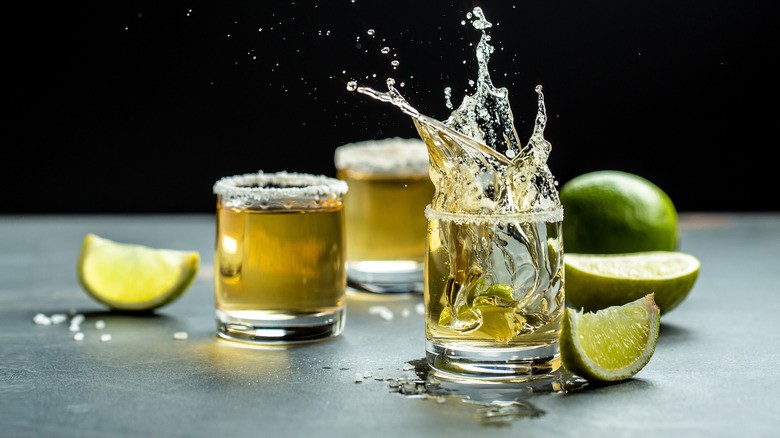 Three glasses of tequila with salt on their rims next to limes; one glass is in mid splash moments after a lime wedge has been dropped inside of it