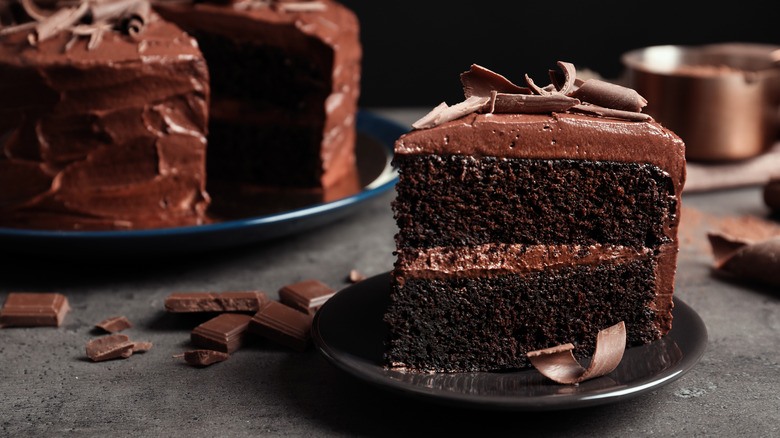 A slice of two layer chocolate cake on a dark gray plate with the rest of the cake in the background