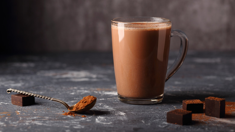 A glass mug of hot cocoa next to a spoon with cocoa powder and pieces of chocolate