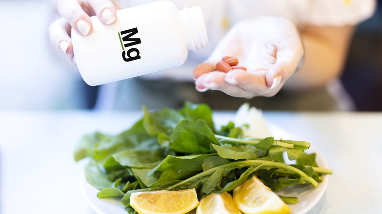 A woman's hand pouring out magnesium supplements before a plate of spinach and oranges