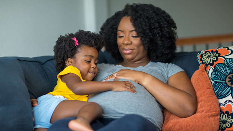 A pregnant mother sits on a blue couch with orange and blue pillows while her toddler daughter puts her hand on her mom's belly. Both daughter and mom are looking down smiling at the mom's belly. 