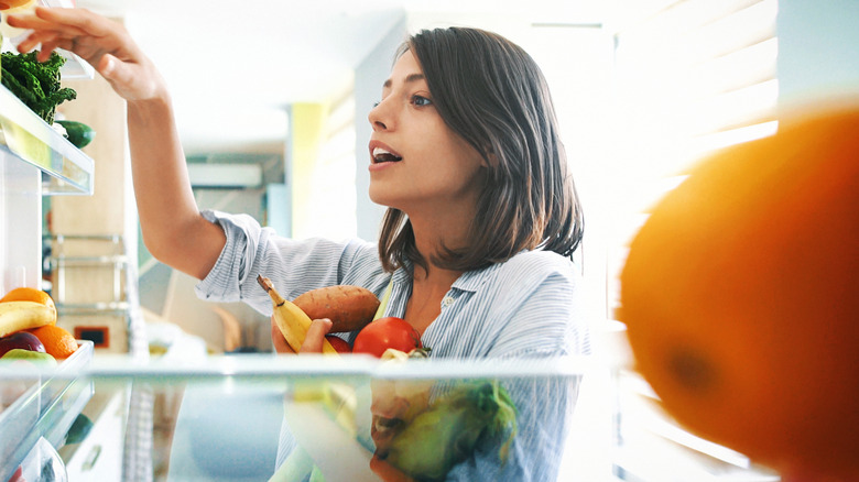 A woman reaching into a refrigerator with an armful of food