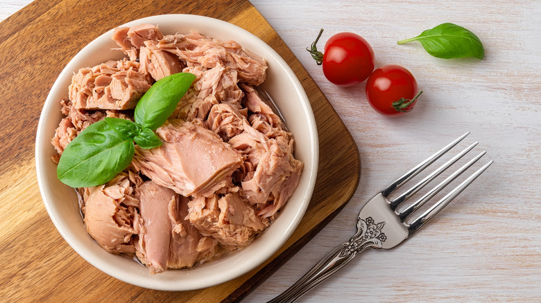 Overhead shot of tuna in a white bowl on a wooden surface next to two small tomatoes and a fork