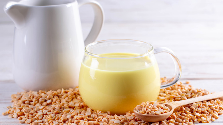 Pea milk in a glass container sitting on top of a pile of yellow peas and next to a wooden spoon and a white pitcher