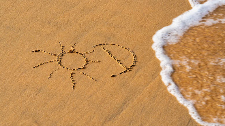 The letter D and a picture of the sun drawn in the sand at the beach next to the surf