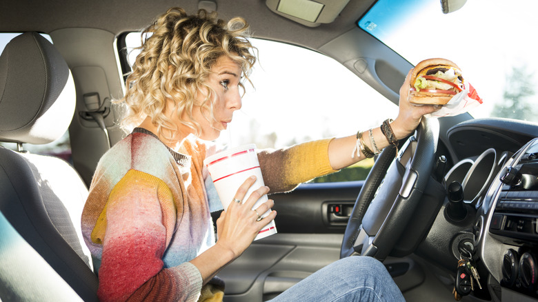 woman drinking a large soft drink in her car while holding a hamburger