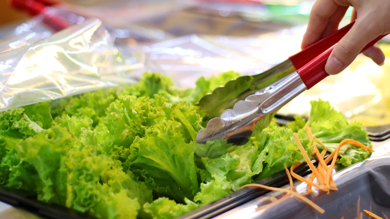 Hand sticking salad tongs into a bed of lettuce at a grocery store salad bar