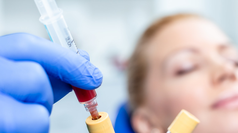 a close up of a syringe and tube filled with blood with a patient in background 