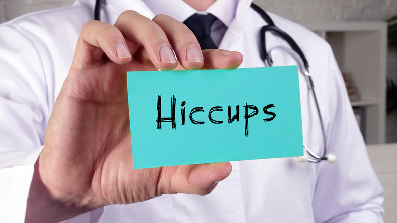 Person in white coat with stethoscope holding a blue green card with the word hiccups written in black