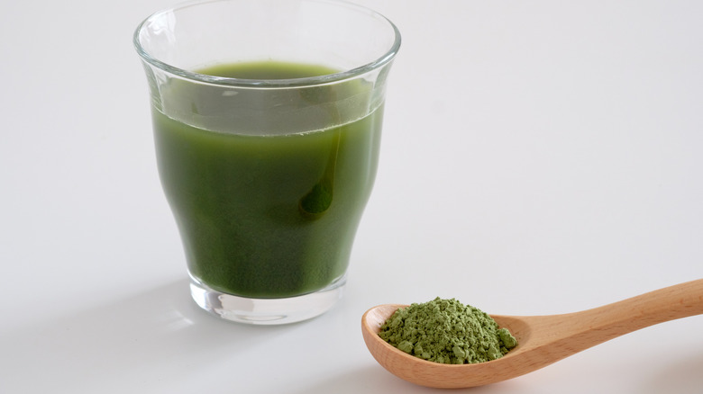 A wooden spoon of greens powder next to a glass of a green drink