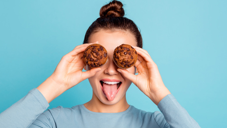 Woman holding chocolate chip muffins over her eyes and sticking out her tongue