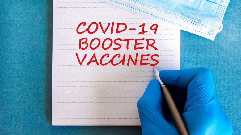 Gloved hand holding a pen to paper writing "COVID-19 BOOSTER VACCINES" in red ink