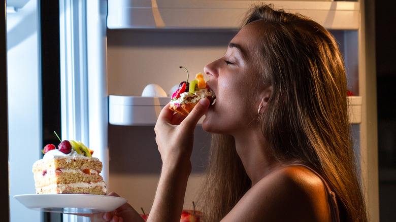 A woman in front of an open fridge eating a tart and holding a large slice of cake