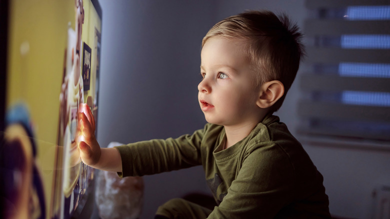 A little boy sitting right in front of a TV and putting his hand on the TV's screen