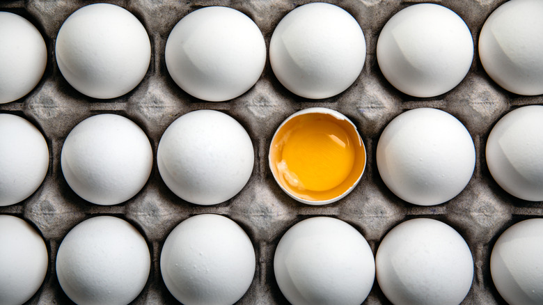 Overhead shot of white eggs in an egg carton, and one egg is broken open, showing it's yolk and egg whites