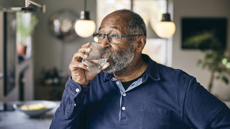 A older man in his kitchen drinking a glass of water.