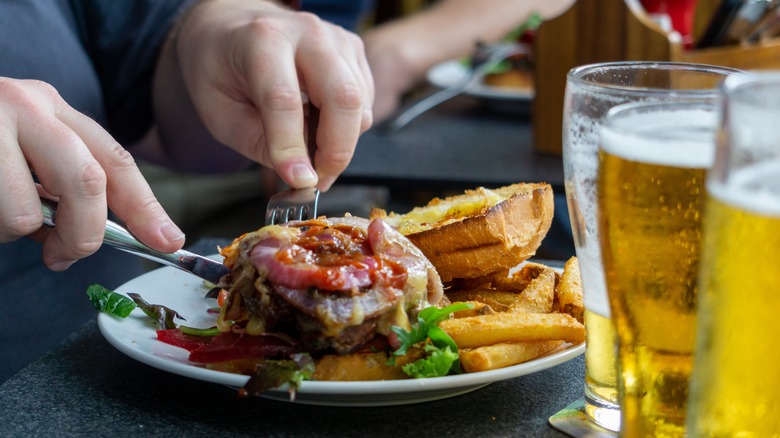 A person eating a cheeseburger and  fries while drinking beer