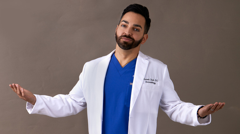 Dr. Muneeb Shah in blue scrubs and a lab coat shrugging