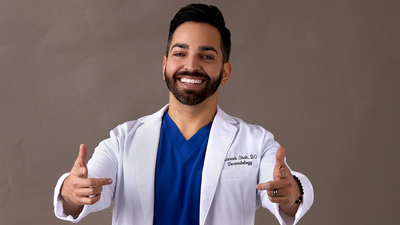 Dr. Muneeb Shah smiling and pointing his fingers at the camera, wearing a lab coat and blue scrubs