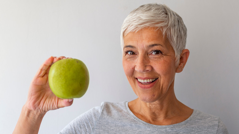 Older woman holding up a green apple and smiling on grey background