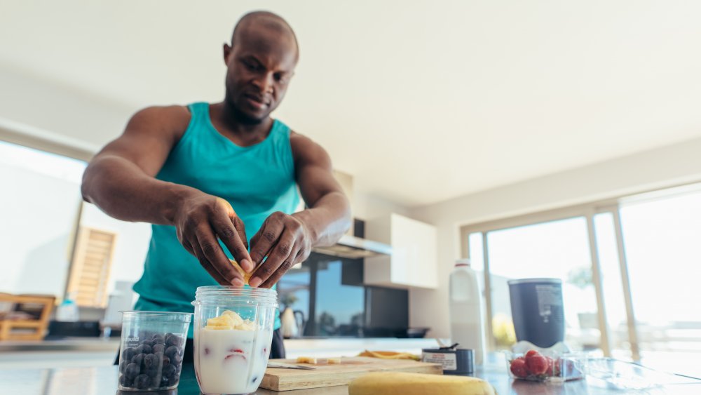 Man preparing milk shake in kitchen. Athletic man adding fruits to jar of milk. Reverse dieting.