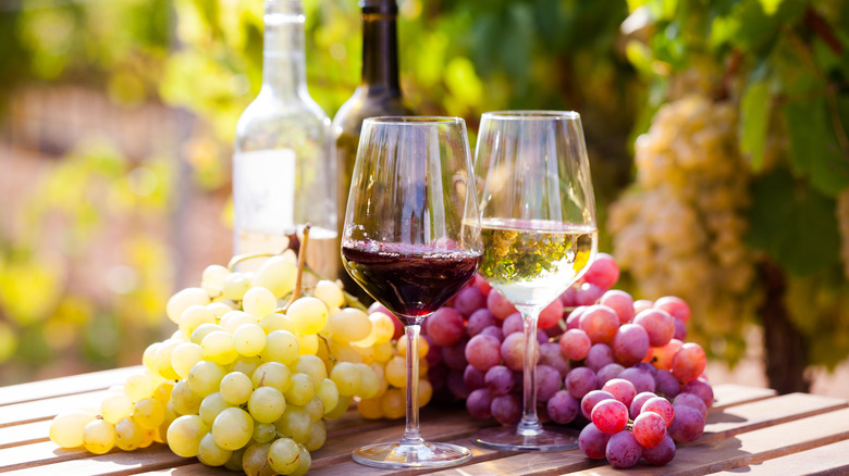 One glass of red wine sitting next to one glass of white wine surrounded by red and green grapes with each wine bottle in the background