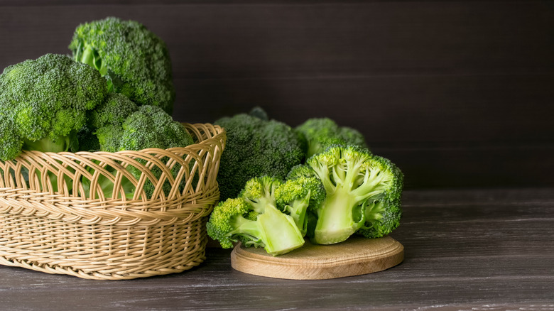 Stalks of Broccoli in a basket next to additional broccoli on a wooden table