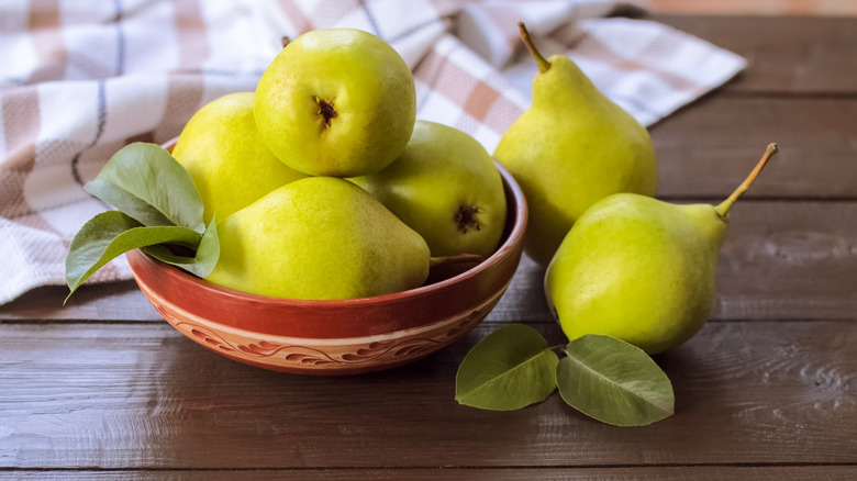 Two green pears next a bowl of pears by a napkin on a wooden table