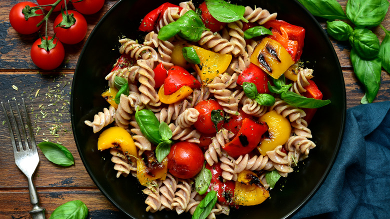 Whole-wheat pasta with basil and vegetables on a black plate next to tomatoes, basil, a blue napkin, and a fork