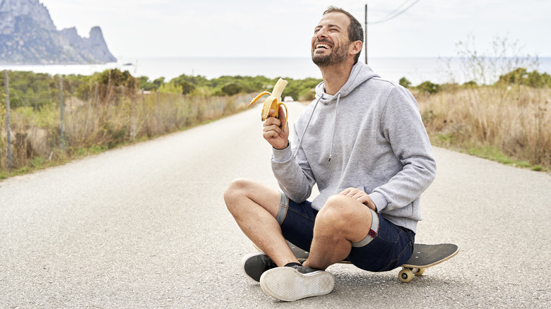 smiling man sitting outside on sidewalk eating banana