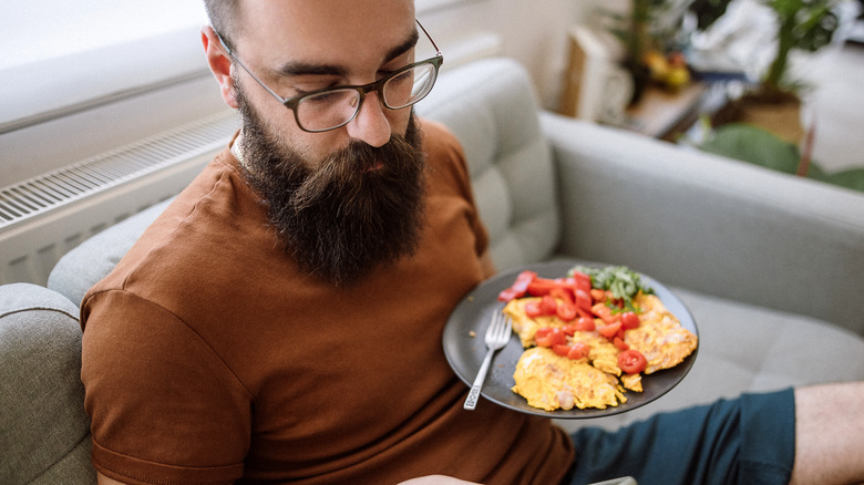 man sitting on sofa eating eggs and other food on plate