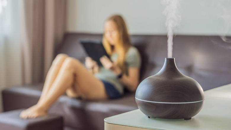 Light haired woman sitting on the couch looking at a tablet and an essential oil diffuser on a table 