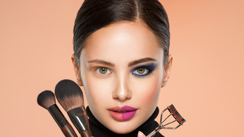 woman holding makeup brushes and eyelash curlers with half of face made up