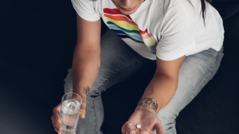 trans woman sitting on a couch, wearing a rainbow shirt holding a pill and a glass of water