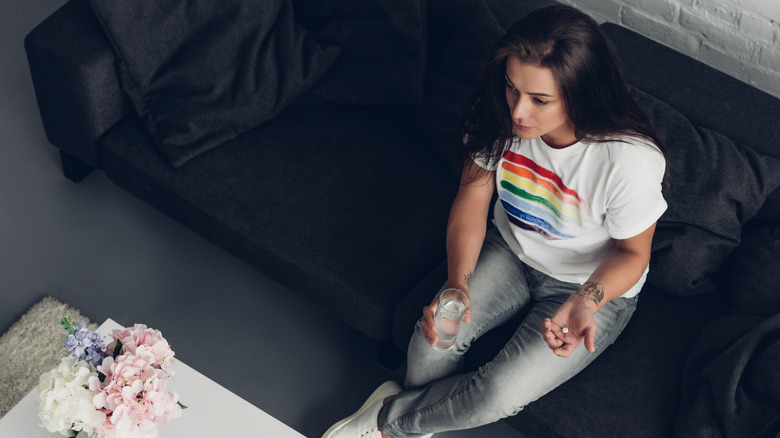 trans woman sitting on a couch, wearing a rainbow flag shirt, holding a glass of water and a pill