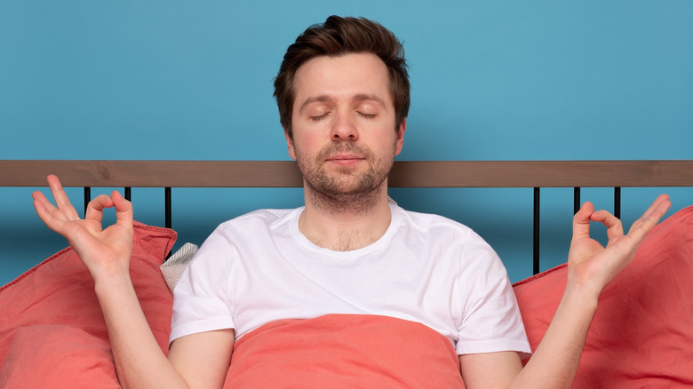 a young man sits in bed with a zen pose 