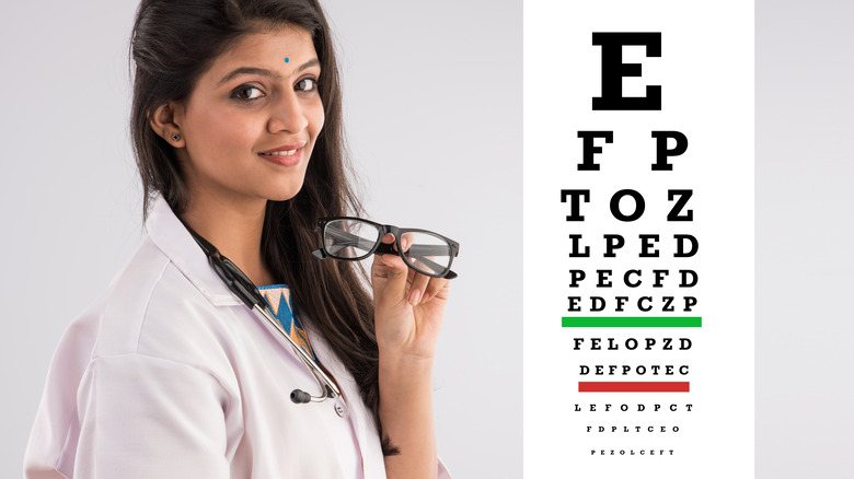 Female ophthalmologist standing next to eye chart and holding glasses