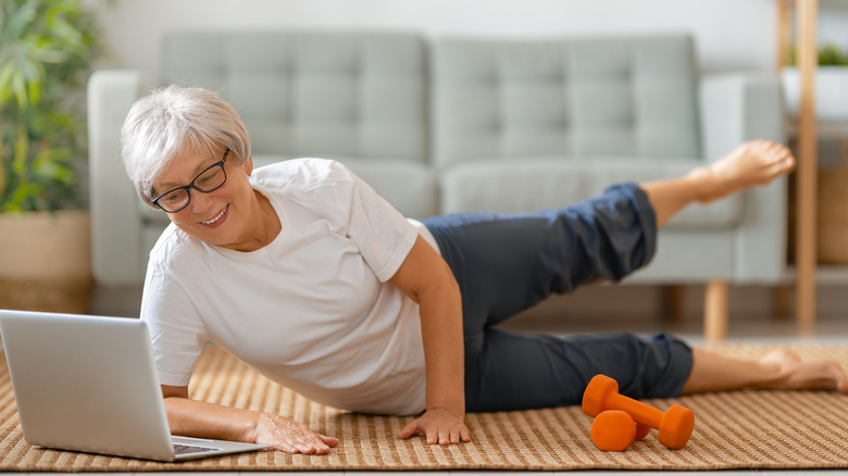 older woman lying on floor doing leg exercises while watching laptop