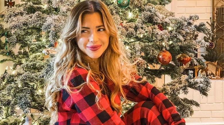Ali Kay sits in front of a Christmas tree in plaid pajamas