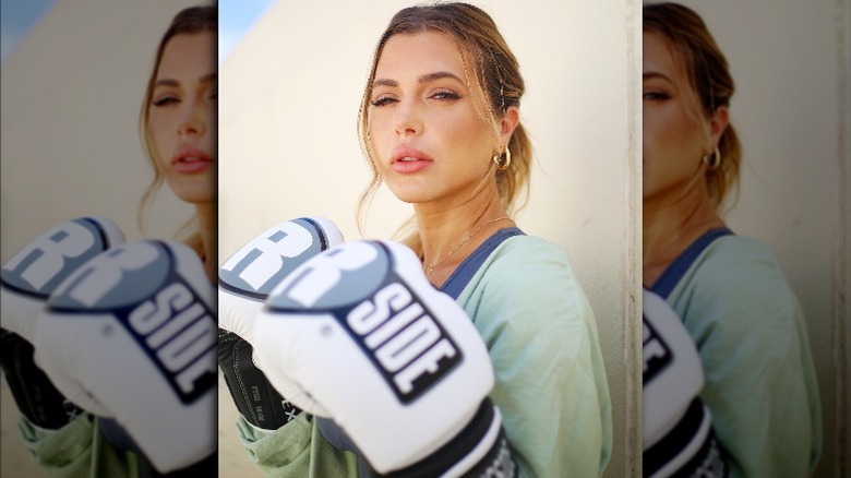 Ali Kay stands against a white wall wearing boxing gloves