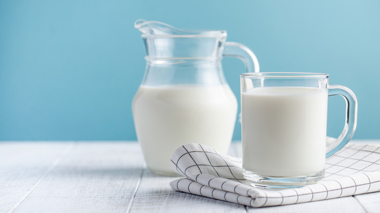 A pitcher of milk next to a glass mug of milk on a napkin 