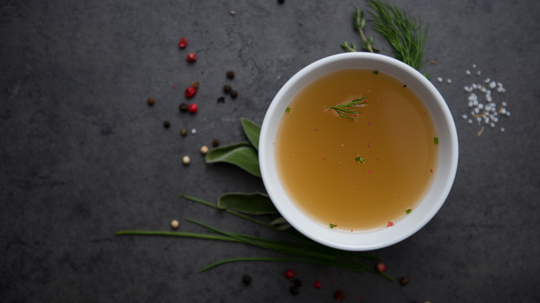 White bowl surrounded by herbs and seasoning and filled with broth