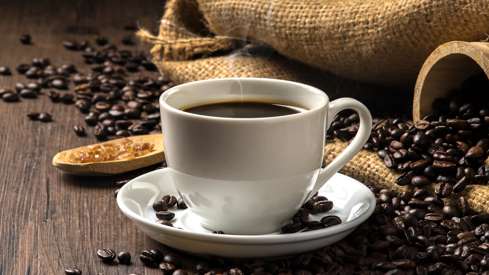 A cup of coffee next to a spilled bag of coffee beans and a spoon full of sugar.